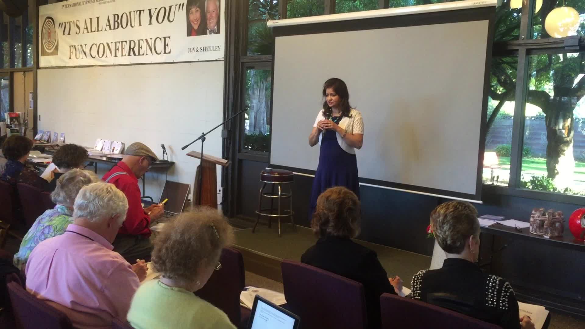 Hena speaking in front of a presentation screen at a live event